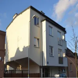 Panorama budynków wielorodzinnych o różnej architekturze i kolorystyce, z balkonami i oknami, w słoneczny dzień. Widoczne elementy otoczenia, takie jak drzewa i ulica.