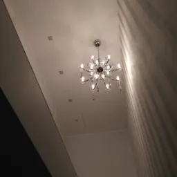 Widok w górę na sufit z zamontowanym ozdobnym, wieloramiennym żyrandolem z żarówkami świecowymi, widoczne także niewielkie kwadratowe oprawy oświetleniowe i fragment ściany z tapetą w pionowe pasy.