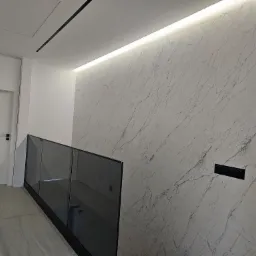 Nowoczesne wnętrze: ściana wykończona płytami imitującymi marmur, podświetlenie LED przy suficie, szklana balustrada schodów, białe drzwi z czarną klamką i czarny panel sterowania na ścianie.