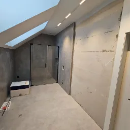 Łazienka w trakcie remontu z kabiną prysznicową w odcieniach szarości, surowa ściana z widocznymi notatkami, oświetlenie punktowe i okno dachowe.