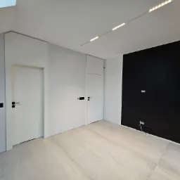 Nowoczesne wnętrze z dwiema białymi drzwiami z czarnymi klamkami, czarną ścianą z obrazem w ramie, oświetleniem LED na suficie i jasną podłogą imitującą marmur.