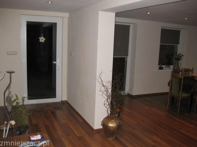 Jasne wnętrze po remoncie z drewnianą podłogą, białymi ścianami i stolarką, widoczne drzwi balkonowe z ozdobą i okna z roletami.