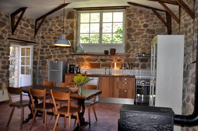 Klimatyczna kuchnia w kamienicy, z drewnianymi belkami stropowymi, kamiennymi ścianami, drewnianymi meblami kuchennymi oraz piecem kaflowym i stołem z krzesłami.