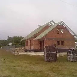 Budowa domu jednorodzinnego w stanie surowym otwartym, widoczne ściany z czerwonej cegły, więźba dachowa, palety z bloczkami oraz ogrodzenie tymczasowe na zewnątrz.