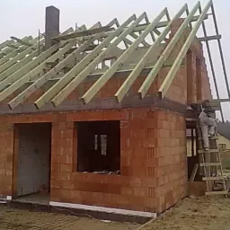 Budowa domu z czerwonej cegły, widoczne ściany, otwory okienne i drewniana konstrukcja dachu w trakcie montażu, pracownik na drabinie.