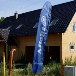 Dom jednorodzinny w trakcie budowy, z widoczną konstrukcją ścian z płyt OSB i czarnym dachem, na pierwszym planie flaga reklamowa dewelopera.