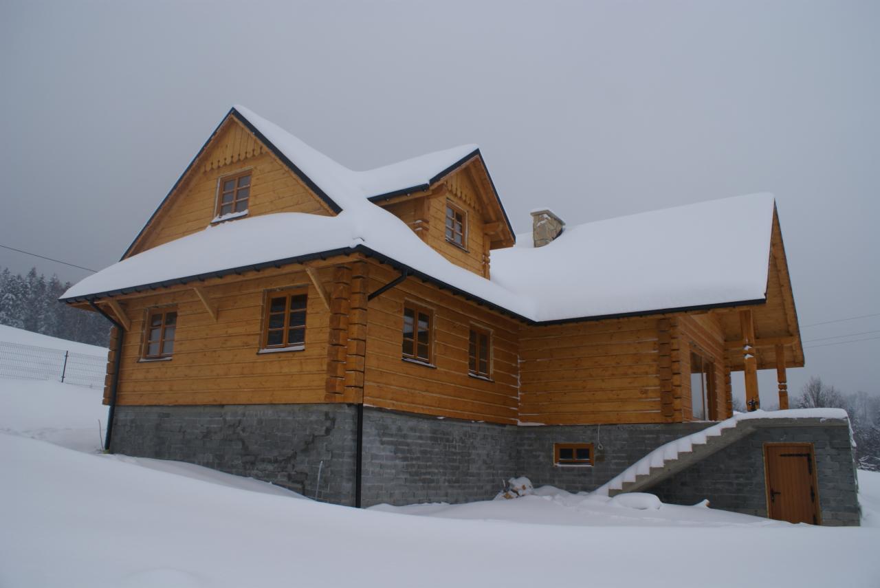 Zimowy krajobraz z widokiem na dwukondygnacyjny dom z bali, częściowo pokryty śniegiem. Dom z kamienną podmurówką, drewnianymi oknami i schodami wejściowymi. Pochmurne niebo w tle.