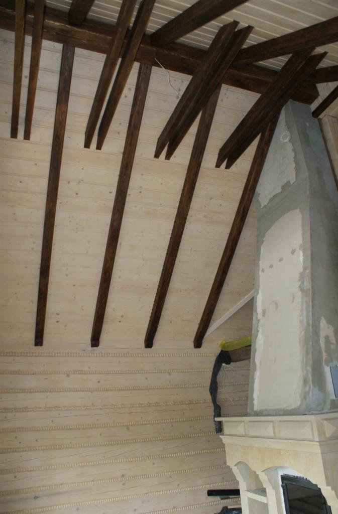 Wnętrze domu z bali: sufit z drewnianych belek w ciemnym kolorze, jasne deski na ścianach z dekoracyjnymi listwami, fragment kominka w trakcie budowy.