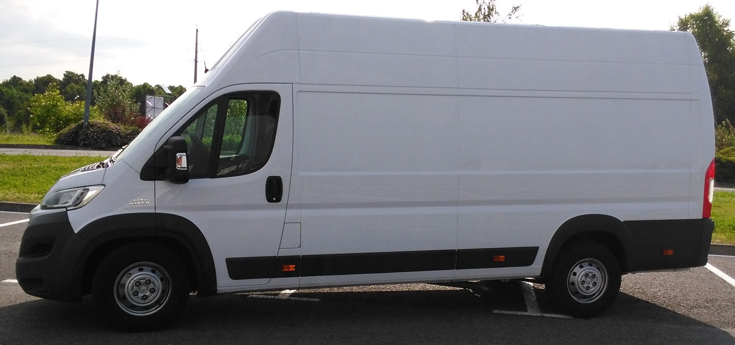 Biały furgon dostawczy z wysokim dachem zaparkowany na parkingu, widok z boku, z zielenią w tle.