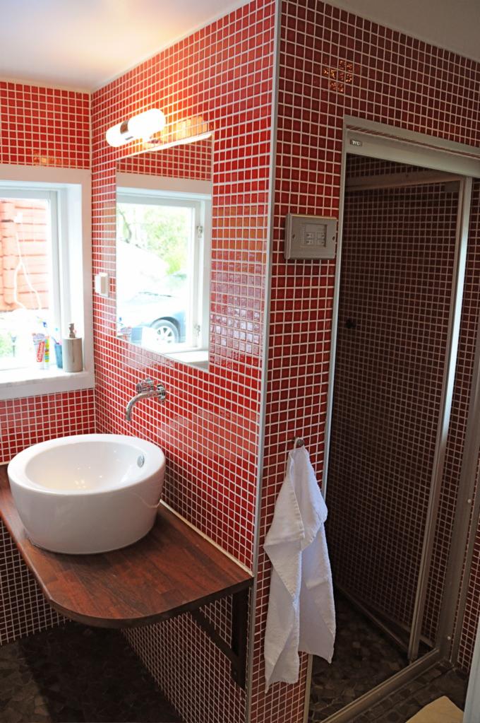 Łazienka wykończona czerwoną mozaiką z białą fugą, z okrągłą białą umywalką na drewnianym blacie i kabiną prysznicową z przesuwnymi drzwiami.