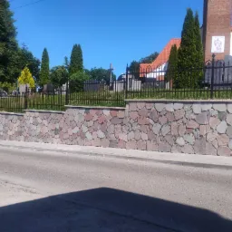 Kamienny murek cmentarny z żelaznym ogrodzeniem w słoneczny dzień. Widoczne nagrobki i zieleń. Estetyczne wykonanie, dbałość o detale.