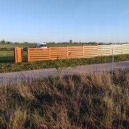 Usługi Ogólnobudowlane Krzysztof Laska - Nowe, drewniane ogrodzenie panelowe z poziomych desek, oddzielające posesję od łąki. W tle biały samochód dostawczy i zieleń. Słoneczny dzień, błękitne niebo.