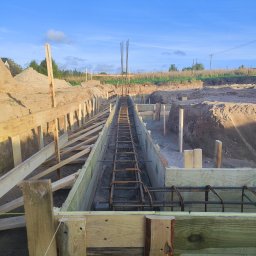 Usługi Ogólnobudowlane Krzysztof Laska - Zbrojenie fundamentów w drewnianym szalunku, widok z góry. Elementy konstrukcyjne i wykop w ziemi. Prace budowlane w toku, niebo w tle.