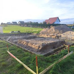 Usługi Ogólnobudowlane Krzysztof Laska - Przygotowanie terenu pod fundamenty: wykop z drewnianym szalunkiem i zbrojeniem, na tle zielonej trawy i zabudowy jednorodzinnej w oddali, w słoneczny dzień.