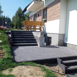 Usługi Ogólnobudowlane Krzysztof Laska - Nowoczesny taras z kostki brukowej w szarym odcieniu, schody prowadzące do domu z drewnianą ławką. Widok na zadbany ogród i elementy placu zabaw w tle.