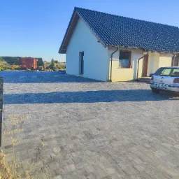 Ułożona kostka brukowa przed domem jednorodzinnym z szarą dachówką, widoczny fragment samochodu osobowego i elementy ogrodu. Słoneczny dzień.