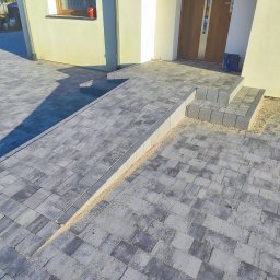 Usługi Ogólnobudowlane Krzysztof Laska - Ułożona szara kostka brukowa przed domem z rampą i schodami z kostki, widoczne gumowce przy drzwiach wejściowych, jasna elewacja.