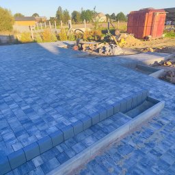 Usługi Ogólnobudowlane Krzysztof Laska - Ułożona, niebiesko-szara kostka brukowa na posesji z widocznymi schodami i elementami przygotowania terenu w tle, w słoneczny dzień.