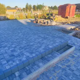 Ułożona, niebiesko-szara kostka brukowa na posesji z widocznymi schodami i elementami przygotowania terenu w tle, w słoneczny dzień.