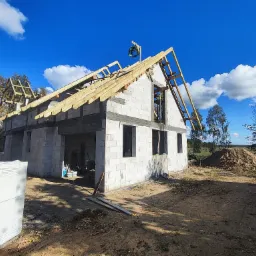 Budowa domu z białych bloczków: surowy stan, więźba dachowa w trakcie montażu, wieniec na szczycie, paleta bloczków obok. Błękitne niebo w tle.