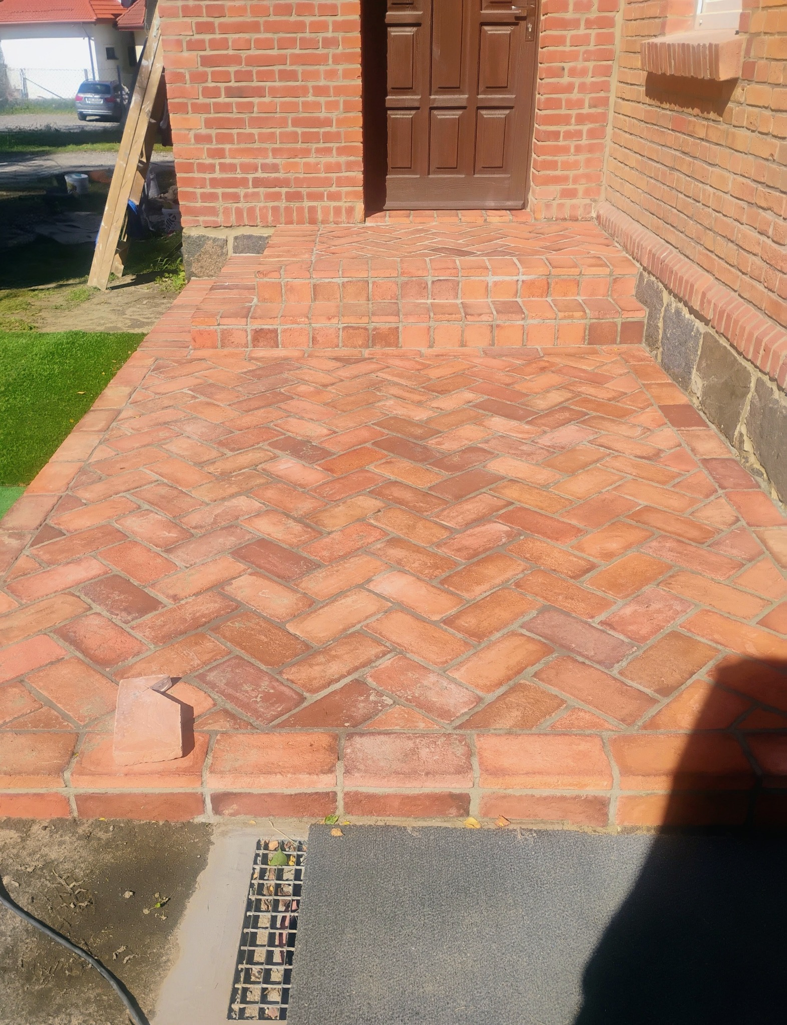 Klinkierowa podłoga tarasu ułożona w jodełkę, prowadząca do drzwi wejściowych. Czerwona cegła na ścianie i schodach tworzy spójny, rustykalny wygląd.