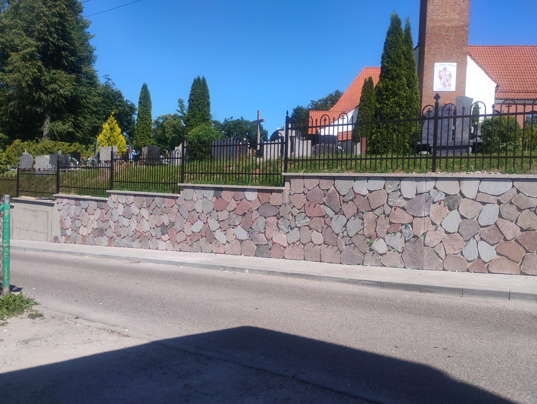 Kamienny murek cmentarny z żelaznym ogrodzeniem w słoneczny dzień. Widoczne nagrobki i zieleń. Estetyczne wykonanie, dbałość o detale.