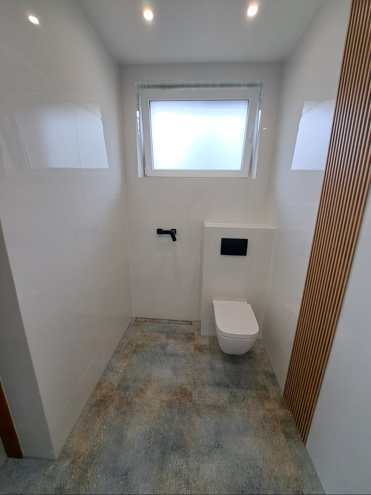 Wykończona łazienka z podwieszaną toaletą, płytkami w odcieniach bieli i szarości oraz drewnianymi panelami ściennymi. Nowoczesny design z oknem.