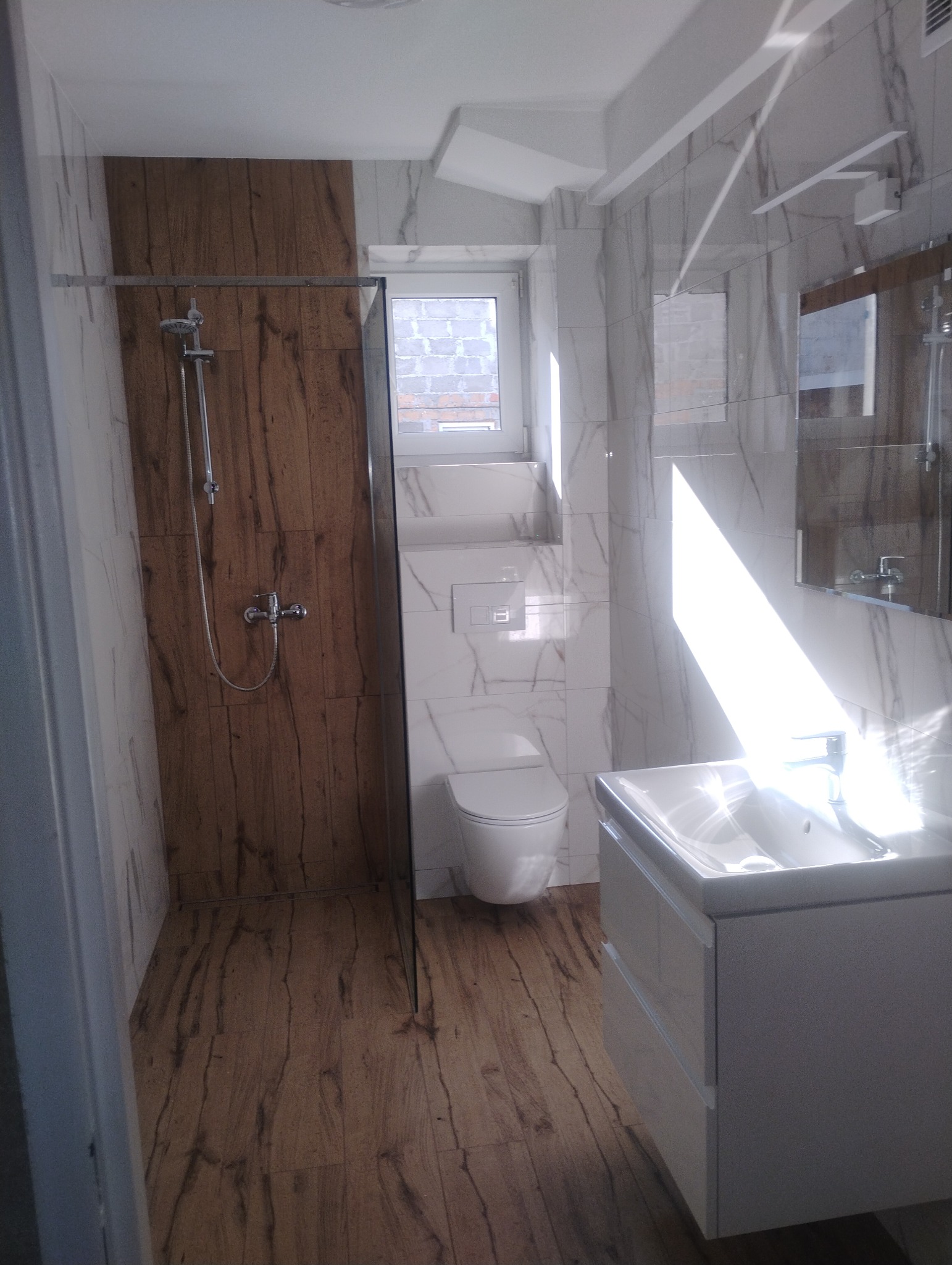 Jasna łazienka po remoncie: kabina prysznicowa z panelami drewnopodobnymi, biała umywalka z szafką i toaleta. Okno z widokiem na budynek.