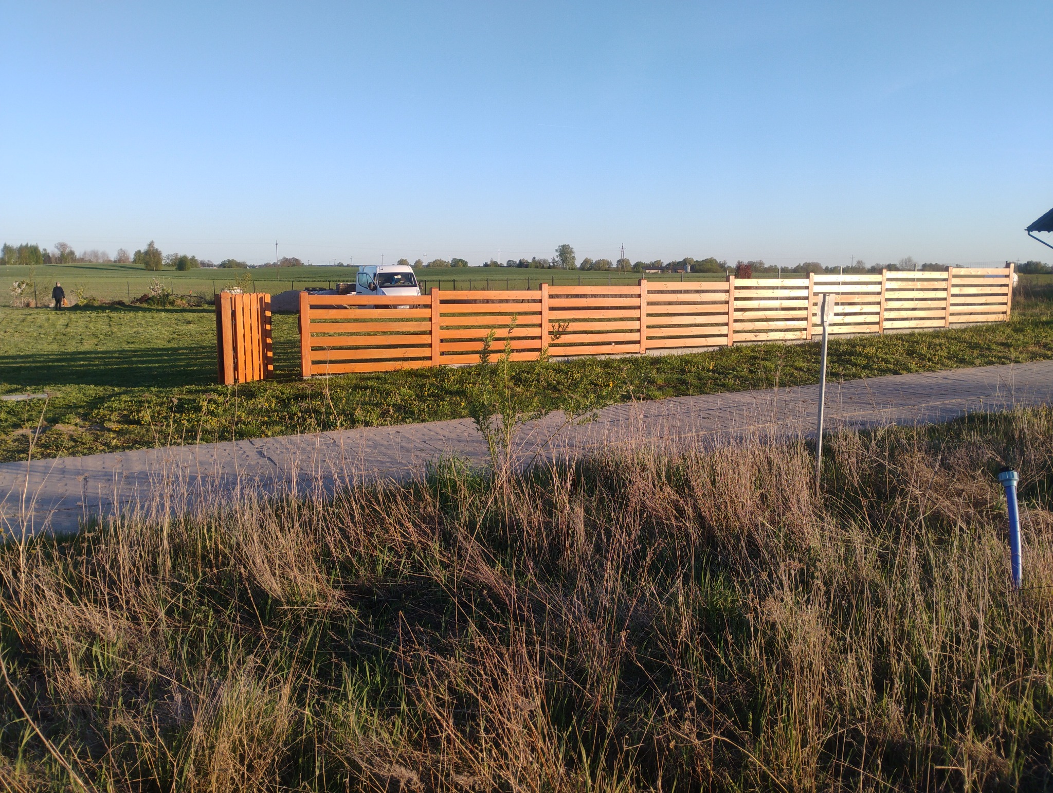 Nowe, drewniane ogrodzenie panelowe z poziomych desek, oddzielające posesję od łąki. W tle biały samochód dostawczy i zieleń. Słoneczny dzień, błękitne niebo.