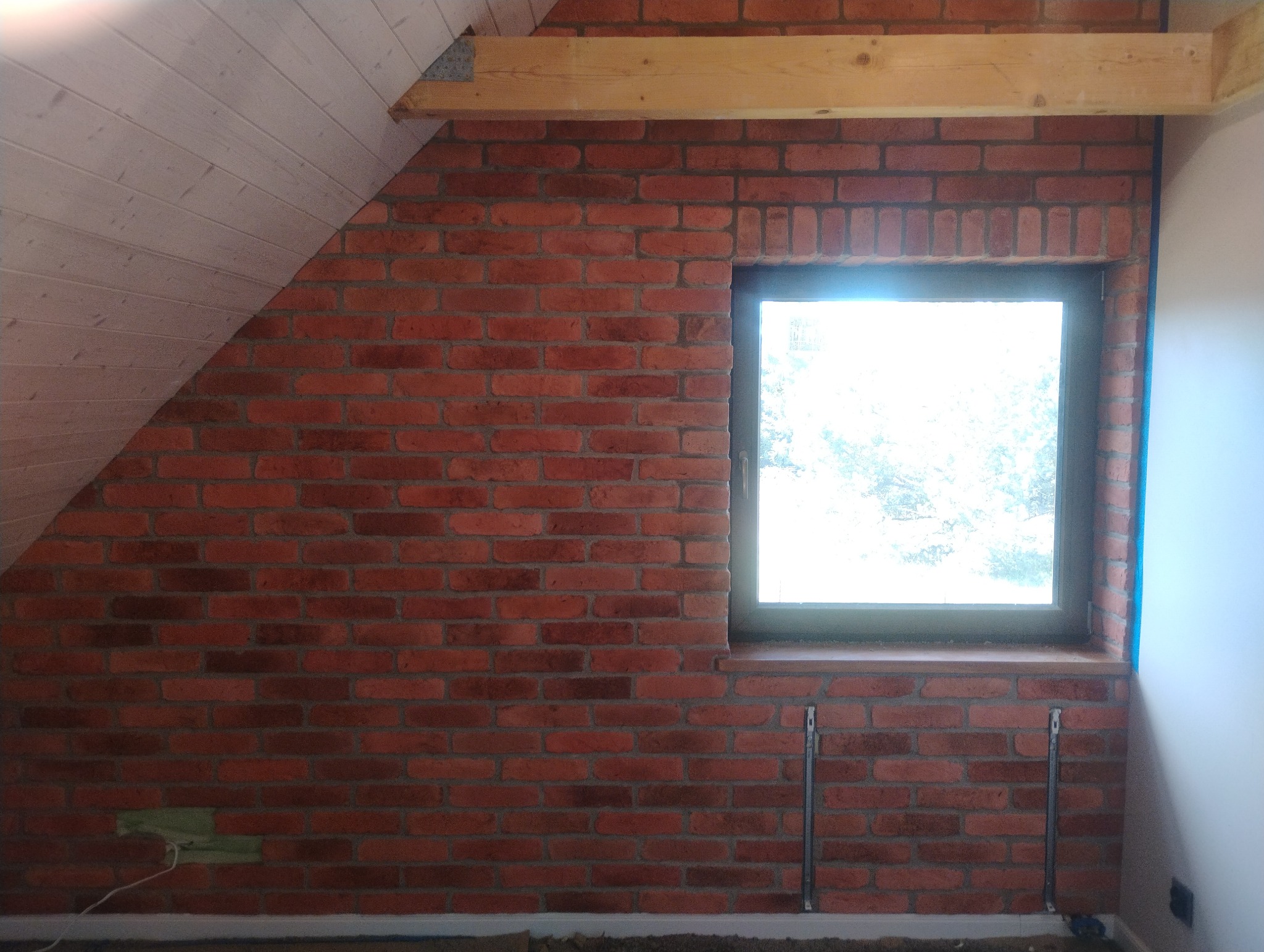 Ceglana ściana z oknem w poddaszu, drewniana belka stropowa, skos z boazerii. Widoczne metalowe wsporniki pod parapetem. Surowy, industrialny styl wykończenia wnętrza.