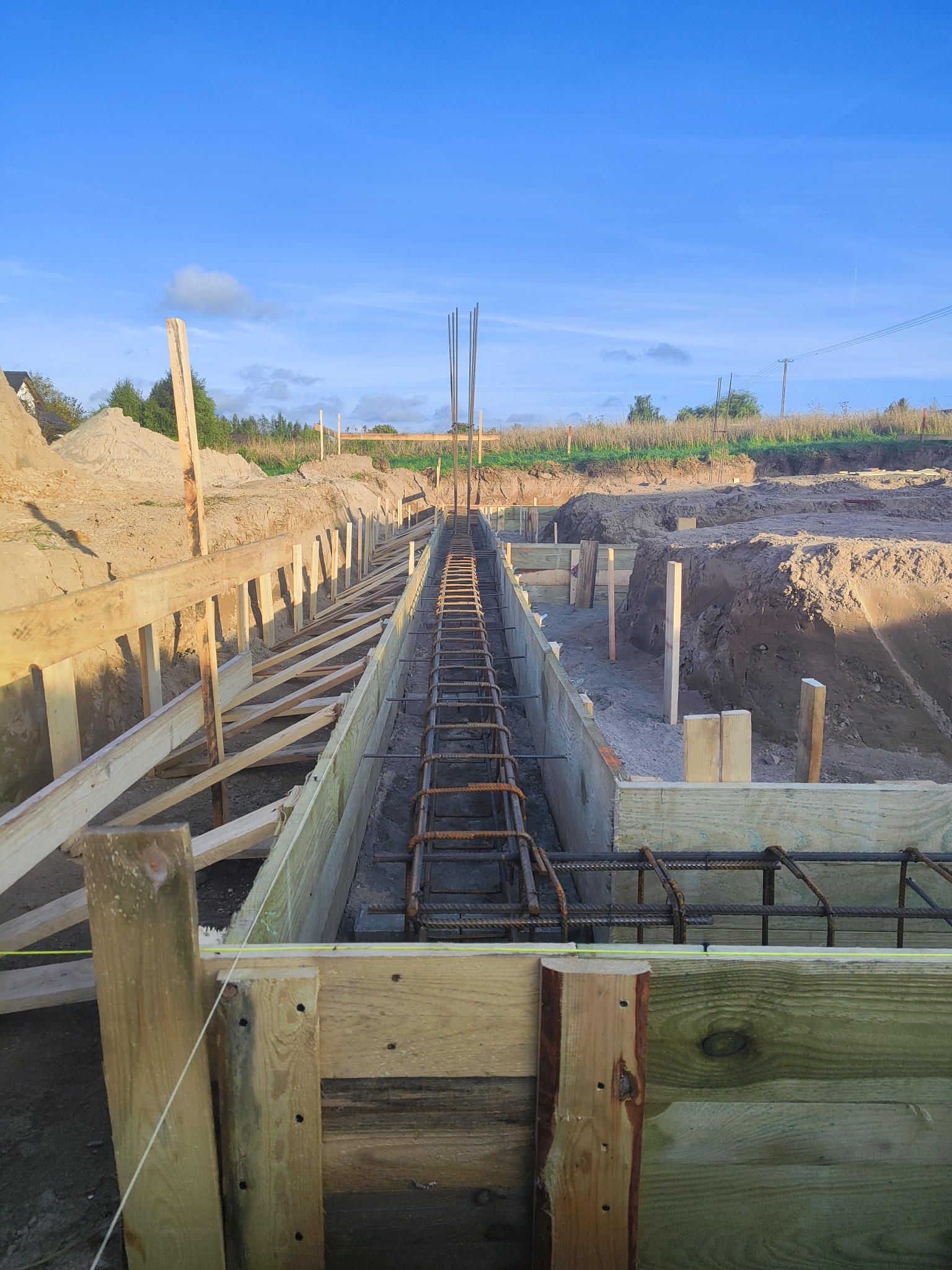 Zbrojenie fundamentów w drewnianym szalunku, widok z góry. Elementy konstrukcyjne i wykop w ziemi. Prace budowlane w toku, niebo w tle.