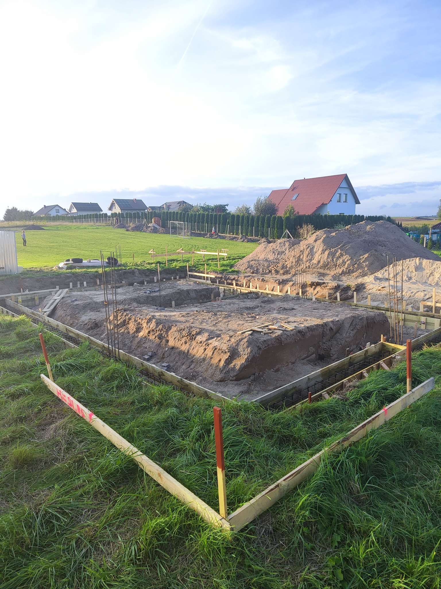 Przygotowanie terenu pod fundamenty: wykop z drewnianym szalunkiem i zbrojeniem, na tle zielonej trawy i zabudowy jednorodzinnej w oddali, w słoneczny dzień.
