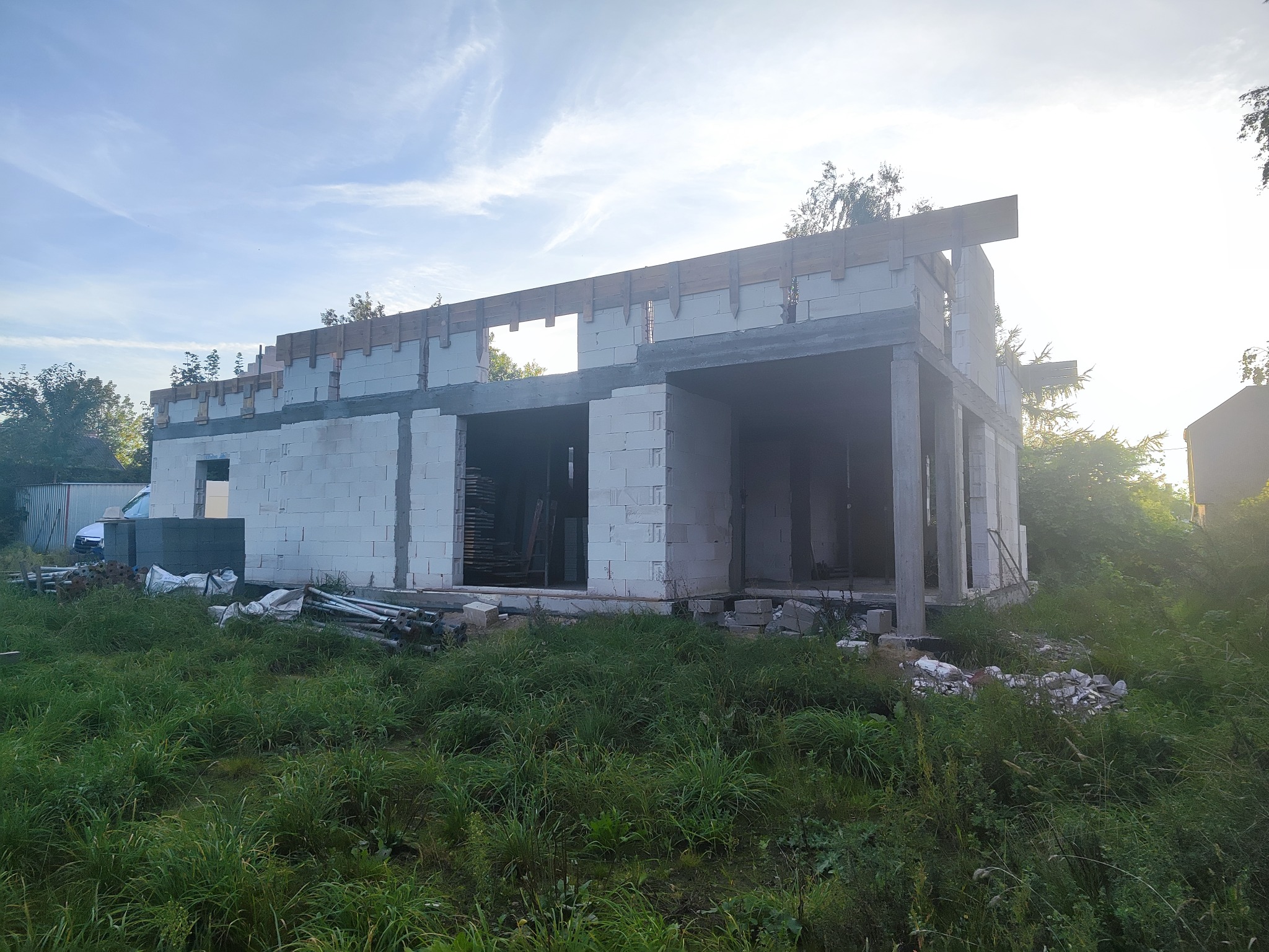 Surowy stan domu z betonu komórkowego, z drewnianymi elementami szalunków, na tle zieleni i błękitnego nieba. Widoczne materiały budowlane i częściowo zarośnięty teren.