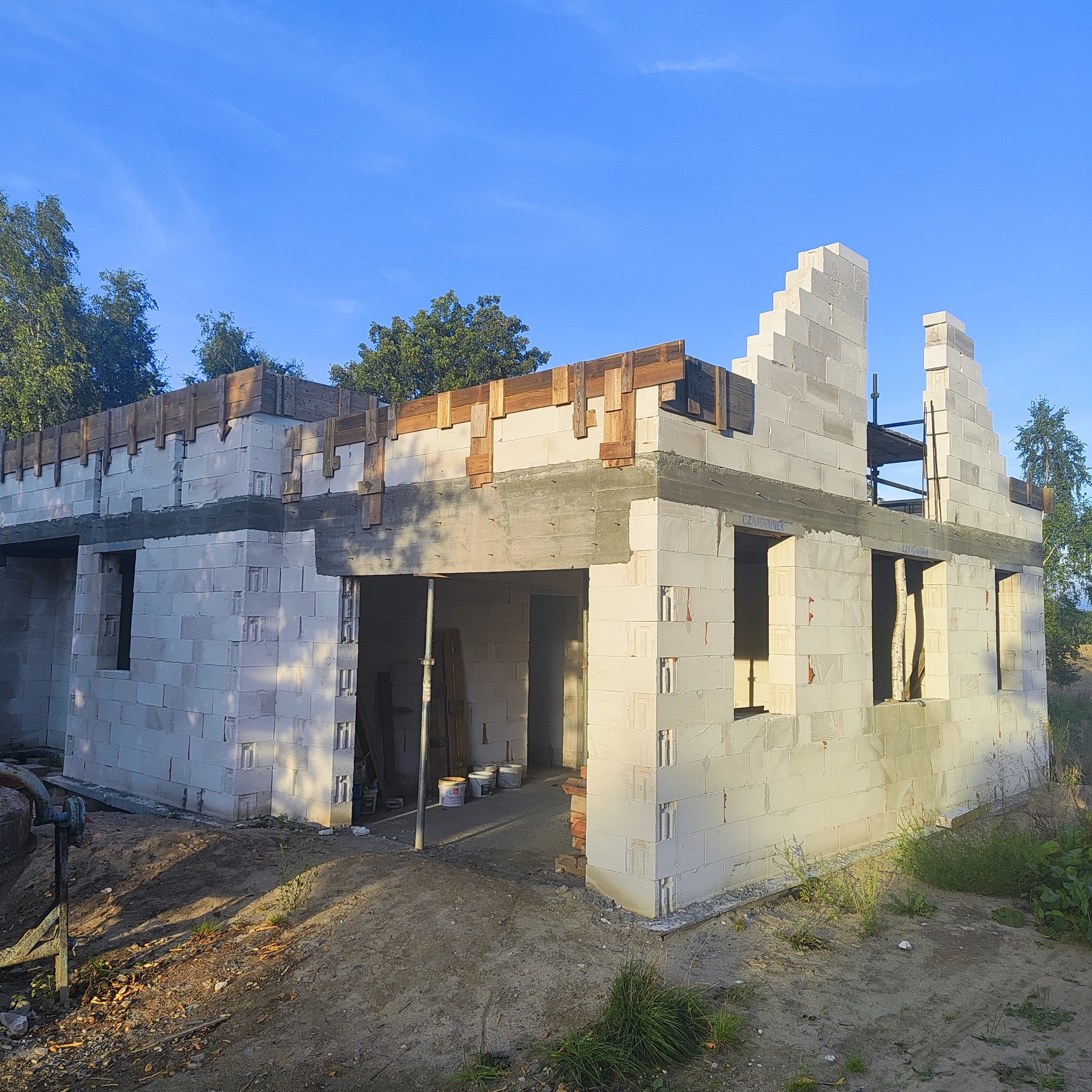 Budowa domu z białych bloczków betonu komórkowego. Widoczne drewniane elementy szalunku na stropie. Częściowo wzniesione ściany, otwory okienne i drzwiowe.
