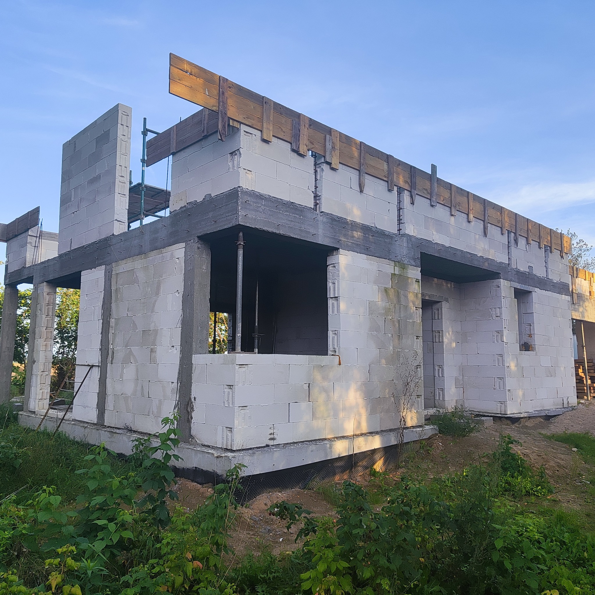 Surowy stan domu murowanego z betonu komórkowego w trakcie budowy. Widoczne szalunki i zbrojenia, fundamenty zabezpieczone, teren zarośnięty. Budowa w toku.