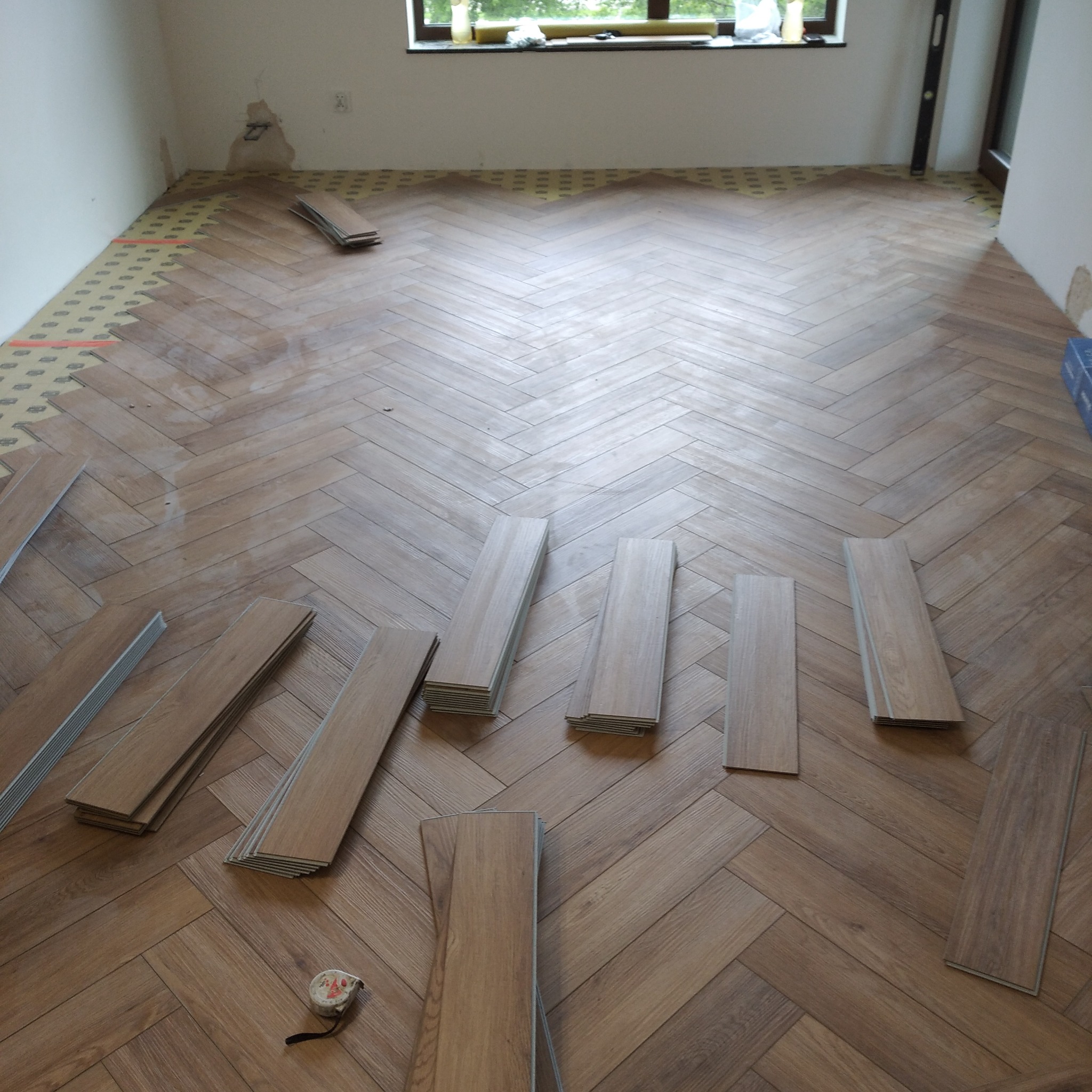 Układanie parkietu w jodełkę: widok na częściowo ułożoną podłogę z paneli drewnianych i narzędzia pomiarowe, z oknem w tle. Etap prac wykończeniowych.