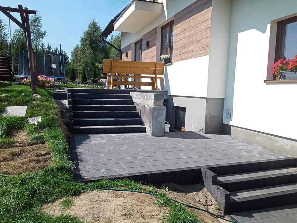 Nowoczesny taras z kostki brukowej w szarym odcieniu, schody prowadzące do domu z drewnianą ławką. Widok na zadbany ogród i elementy placu zabaw w tle.