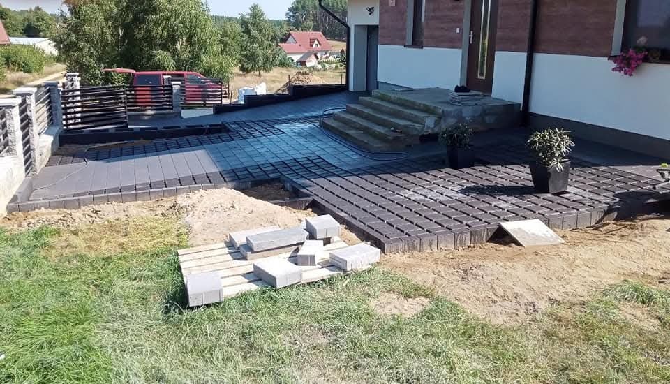 Układanie szarej kostki brukowej na podjeździe i tarasie domu jednorodzinnego. Widoczne niezakończone prace i przygotowane materiały.