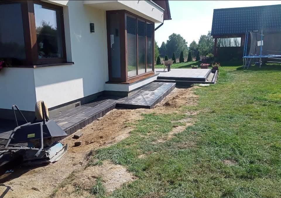 Nowoczesny taras z kamienną nawierzchnią przy domu z dużymi oknami, w tle zielony trawnik i budynek gospodarczy z trampoliną. Widoczny częściowo sprzęt budowlany.