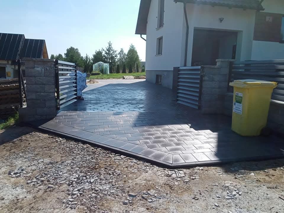 Nowa, szara kostka brukowa ułożona na podjeździe przed domem, z widoczną bramą wjazdową i żółtym pojemnikiem na śmieci. Jasne, słoneczne otoczenie.