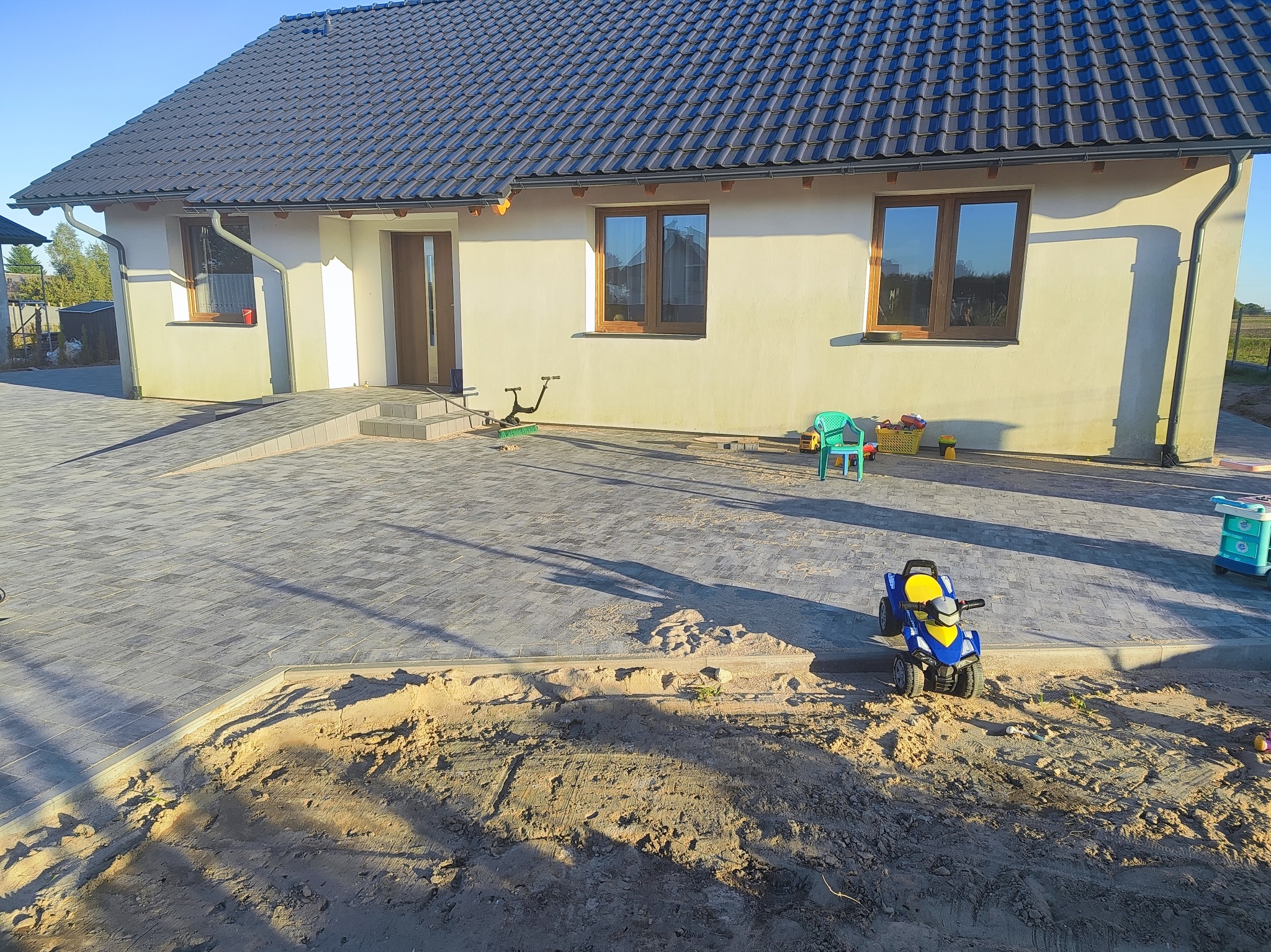 Nowoczesny dom z ciemnym dachem i jasną elewacją. Ułożona kostka brukowa na podjeździe, widoczne elementy placu zabaw. Słoneczny dzień.