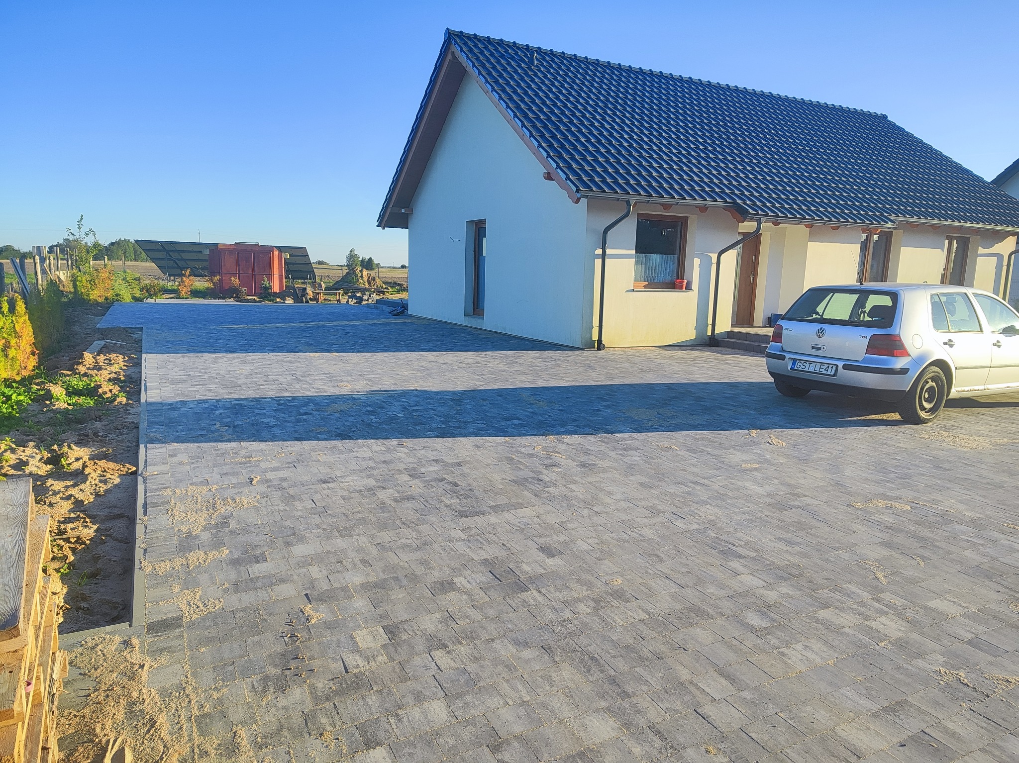 Ułożona kostka brukowa przed domem jednorodzinnym z szarą dachówką, widoczny fragment samochodu osobowego i elementy ogrodu. Słoneczny dzień.