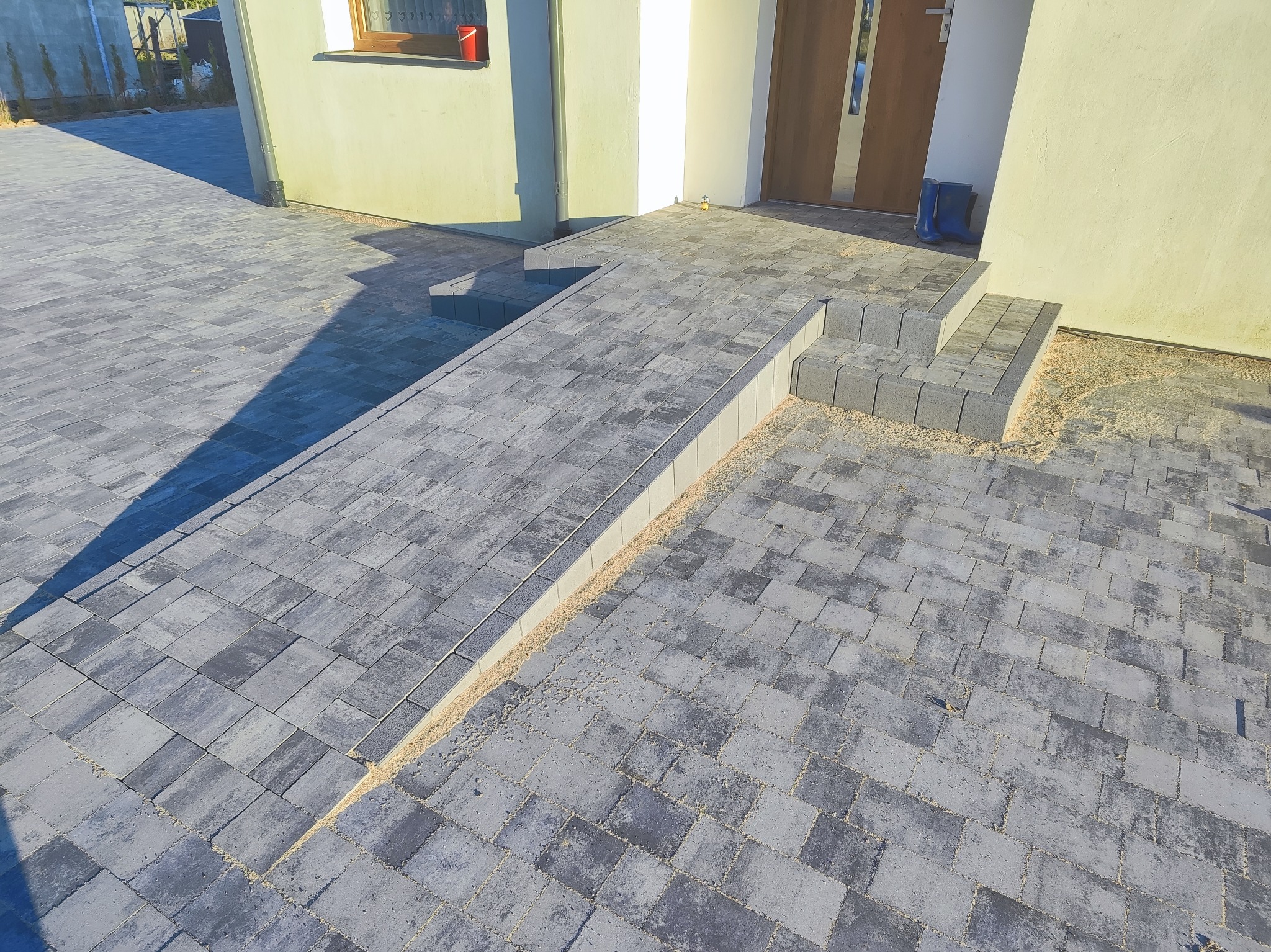Ułożona szara kostka brukowa przed domem z rampą i schodami z kostki, widoczne gumowce przy drzwiach wejściowych, jasna elewacja.