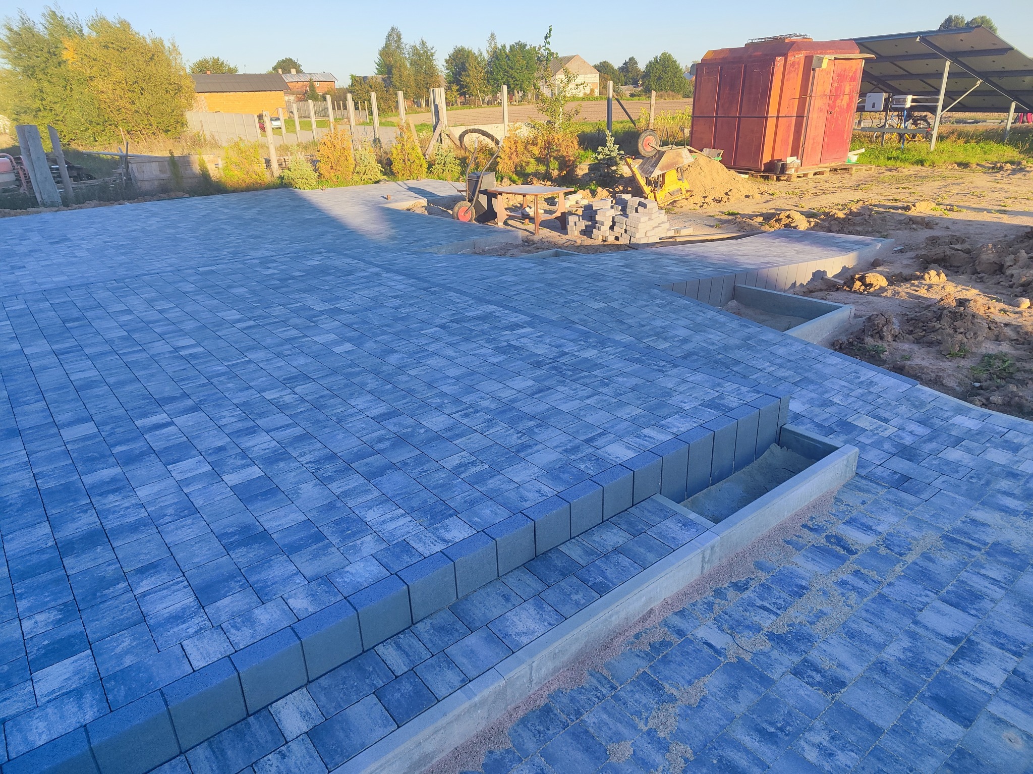 Ułożona, niebiesko-szara kostka brukowa na posesji z widocznymi schodami i elementami przygotowania terenu w tle, w słoneczny dzień.