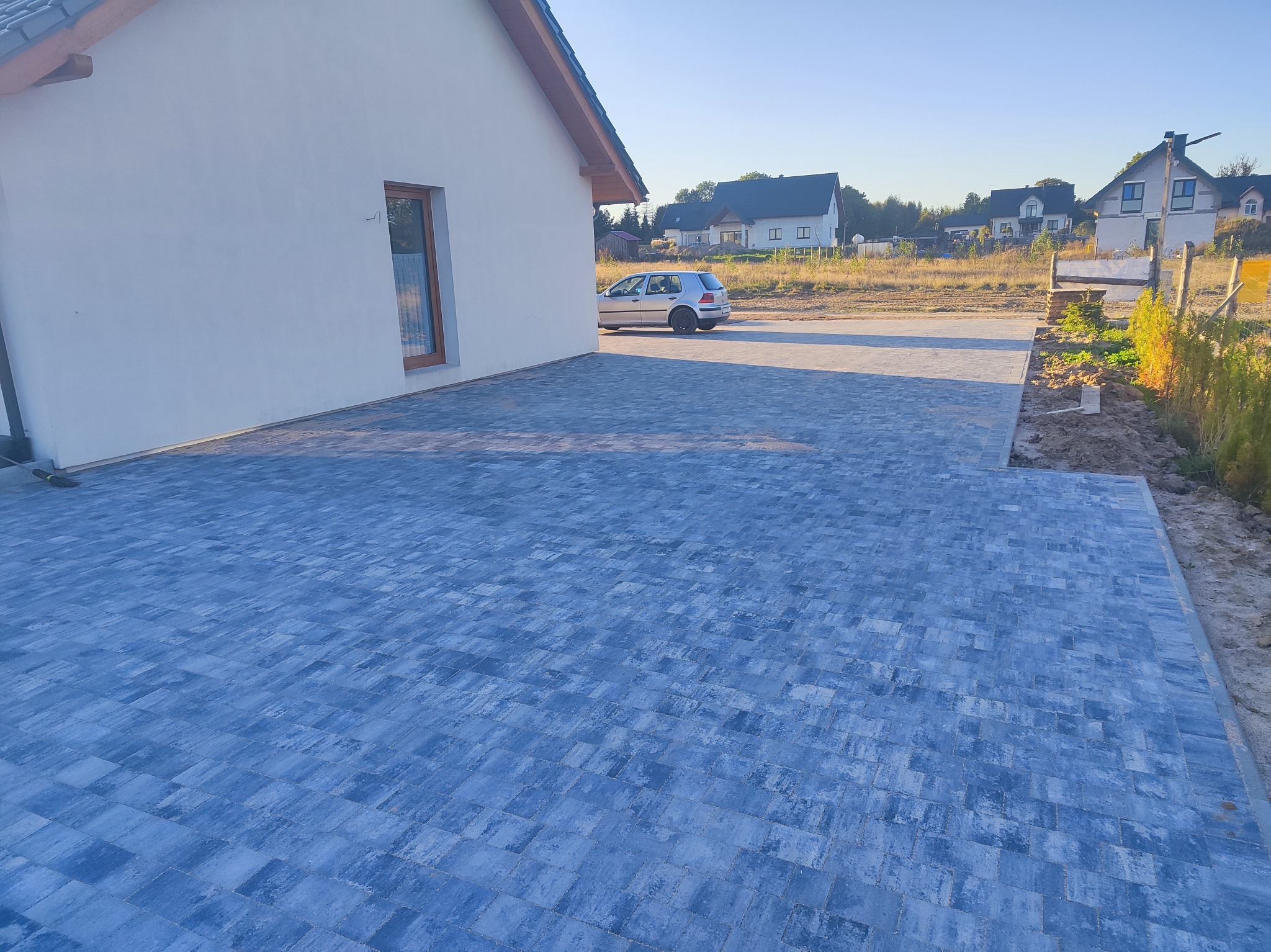 Ułożona, szara kostka brukowa na podjeździe przed domem jednorodzinnym. W tle zaparkowany samochód osobowy i inne domy w oddali, w słoneczny dzień.