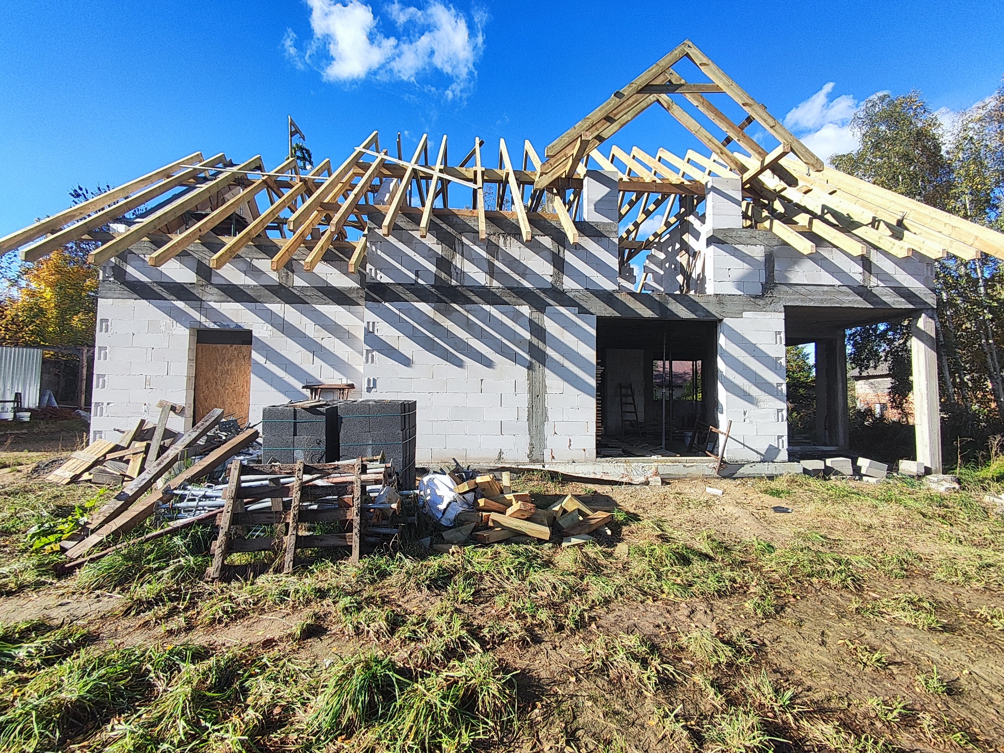 Surowy stan domu z bloczków, widoczne drewniane elementy konstrukcji dachu, resztki materiałów budowlanych na trawiastym terenie. Niebieskie niebo w tle.