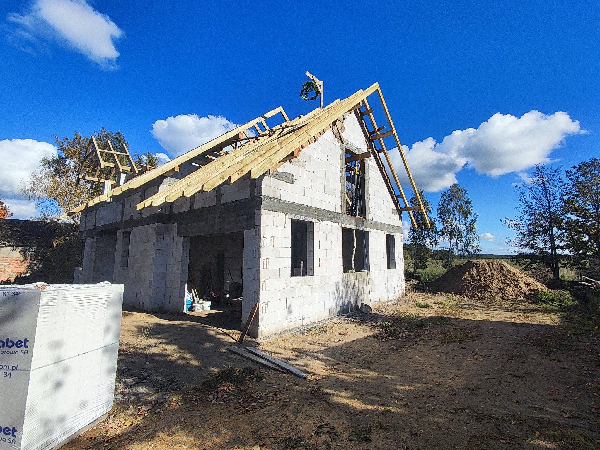 Budowa domu z białych bloczków: surowy stan, więźba dachowa w trakcie montażu, wieniec na szczycie, paleta bloczków obok. Błękitne niebo w tle.