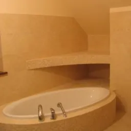 Owalna wanna obudowana mozaiką w beżowym odcieniu, dopasowana kolorystycznie do płytek ściennych i półki wnękowej, chromowana armatura, minimalistyczny wystrój łazienki.