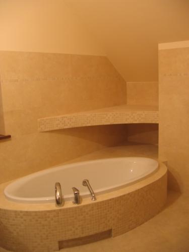 Owalna wanna obudowana mozaiką w beżowym odcieniu, dopasowana kolorystycznie do płytek ściennych i półki wnękowej, chromowana armatura, minimalistyczny wystrój łazienki.