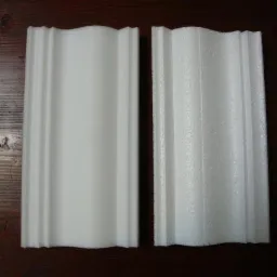 Dwa białe elementy dekoracyjne z tworzywa sztucznego o falistym kształcie, jeden gładki, drugi o porowatej strukturze, leżące na ciemnym, drewnianym tle.